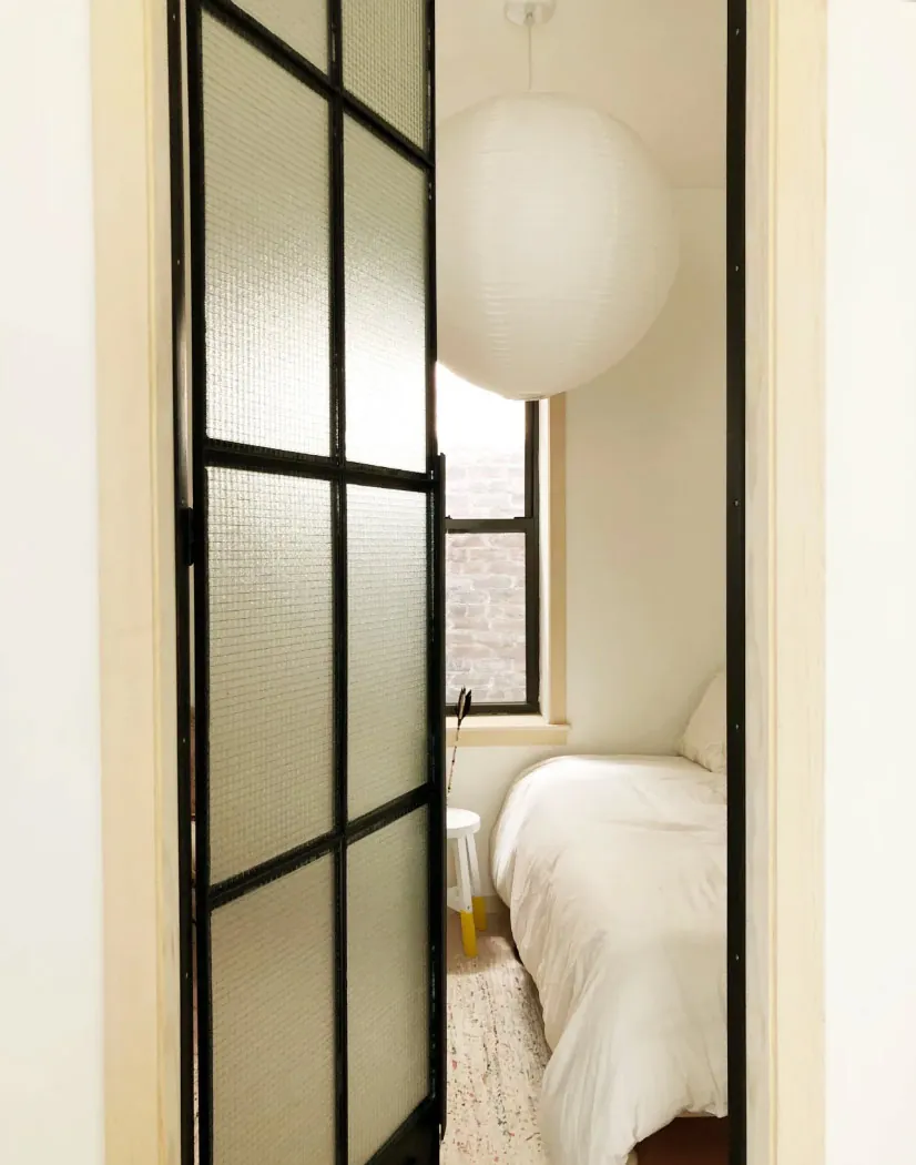 Second bedroom with a paper globe pendant light behind a black metal framed glass door in a renovated NoHo apartment