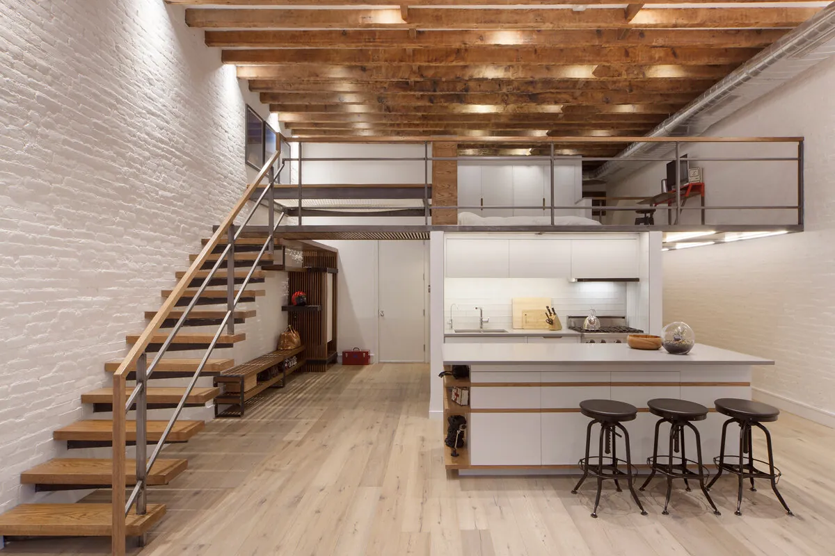 Open Soho loft in New York with exposed wood beams, white brick walls, a staircase and mezzanine above the kitchen