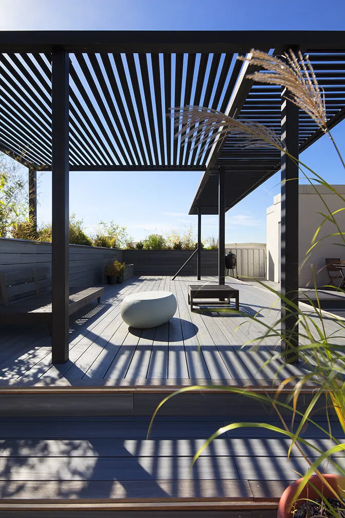 Rooftop deck under a slatted steel pergola with built-in bench seating and planters by MELDNYC