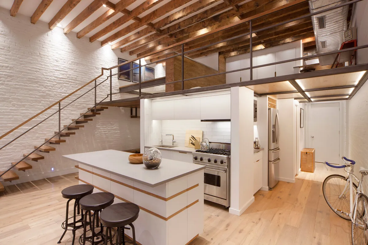 Soho kitchen in New York with white cabinetry, a wood-trimmed island and three dark stools beneath a mezzanine