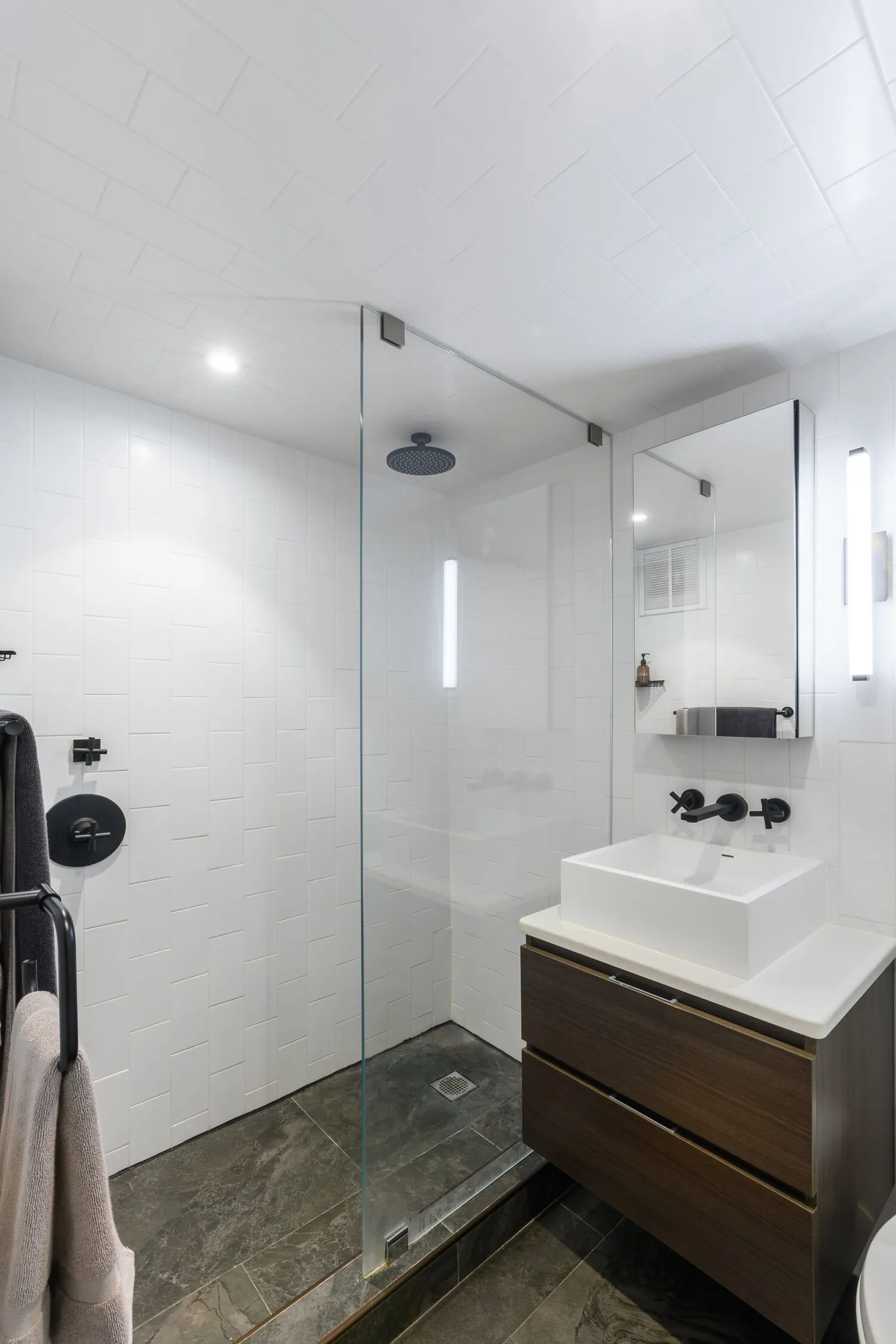 Lexington bathroom in New York City with white tiled walls, a glass shower, wood vanity and black wall-mounted fixtures