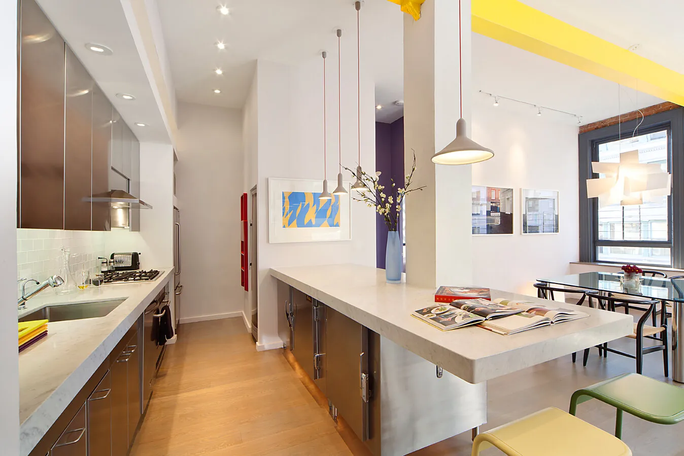 Galley kitchen in a Greenwich Village apartment renovation with a long white island, pendant lights and a yellow accent wall